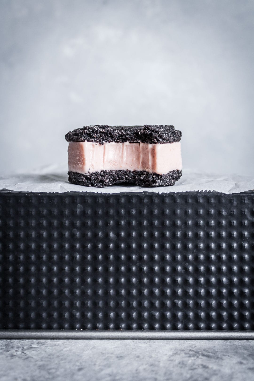 Side view of chocolate blood orange ice cream sandwich with bite taken out, resting on parchment paper and a black metal tin