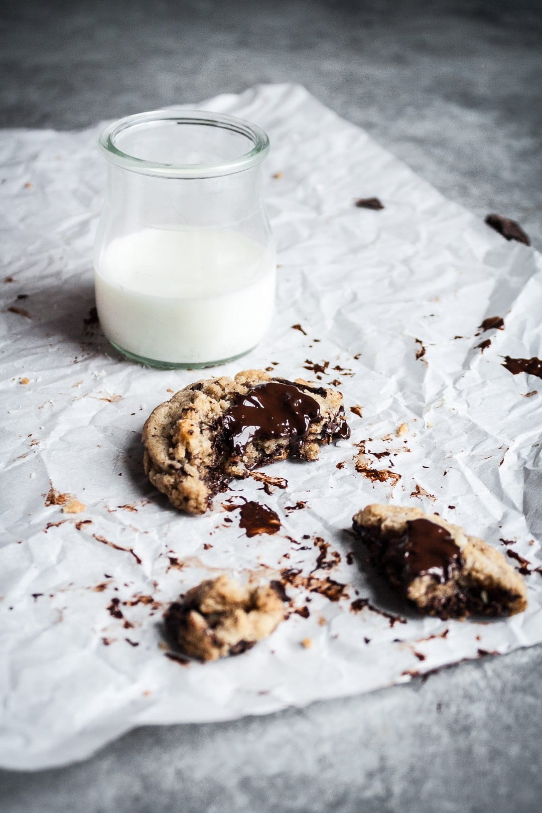 A partially eaten coconut chocolate chip cookie on parchment paper with a glass of milk nearby