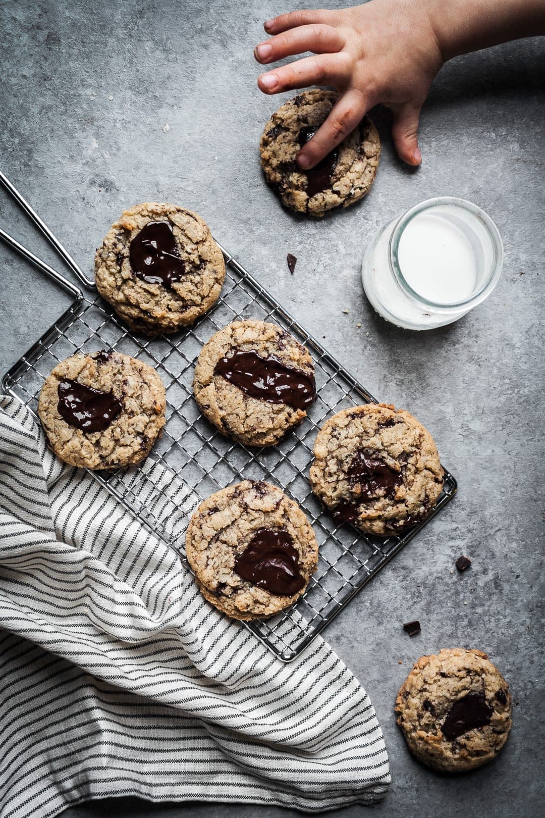 Freshly baked cookies on a cooling rack with napkin, glass of milk on a grey background with a toddler's hands reaching in to touch the cookie