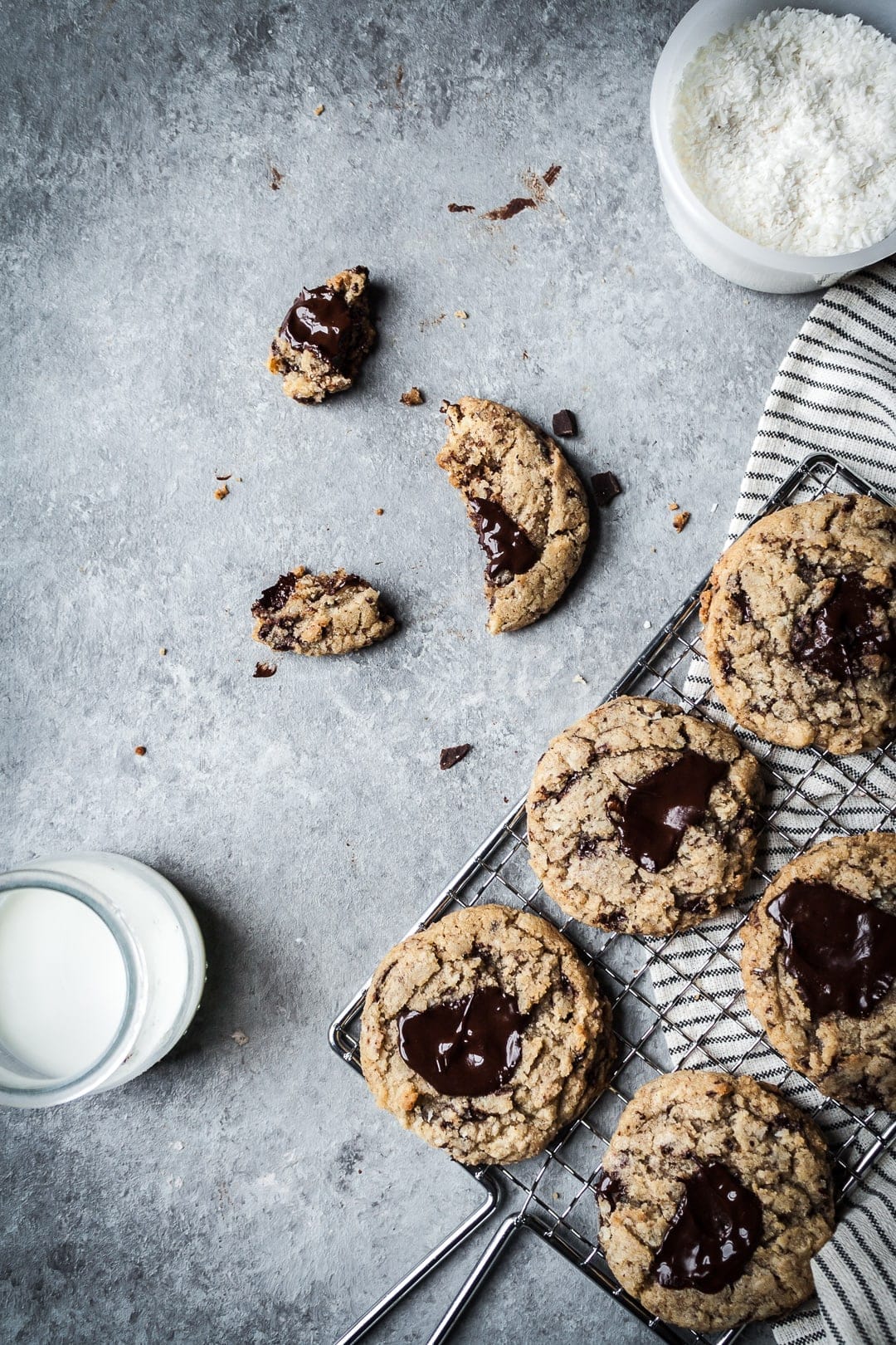 Freshly baked cookies on a cooling rack with a partially eaten cookie nearby, along with a glass of milk and a bowl of shredded coconut