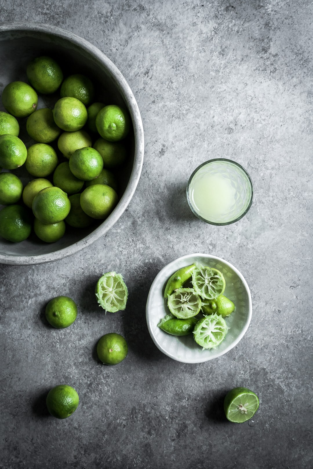 Key limes in a bowl, some cut in half and squeezed, with a glass of key lime juice on a grey background