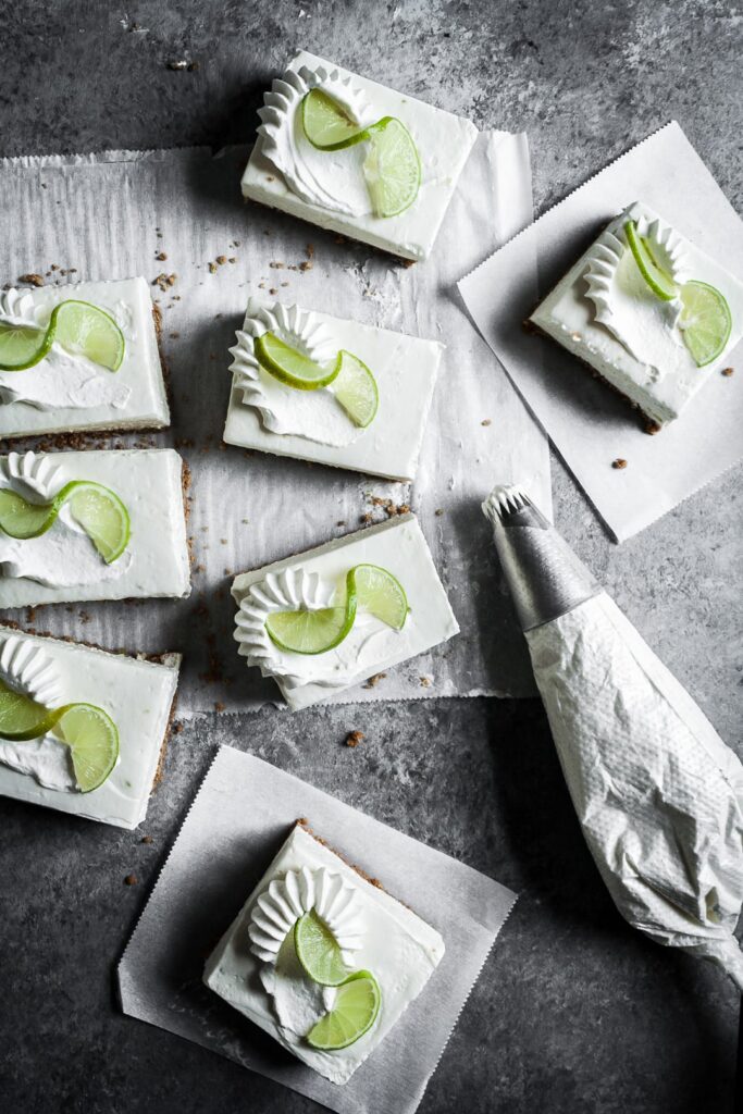 Top view of sliced cheesecake bars on parchment paper squares on a grey background with a piping bag of whipped cream nearby.