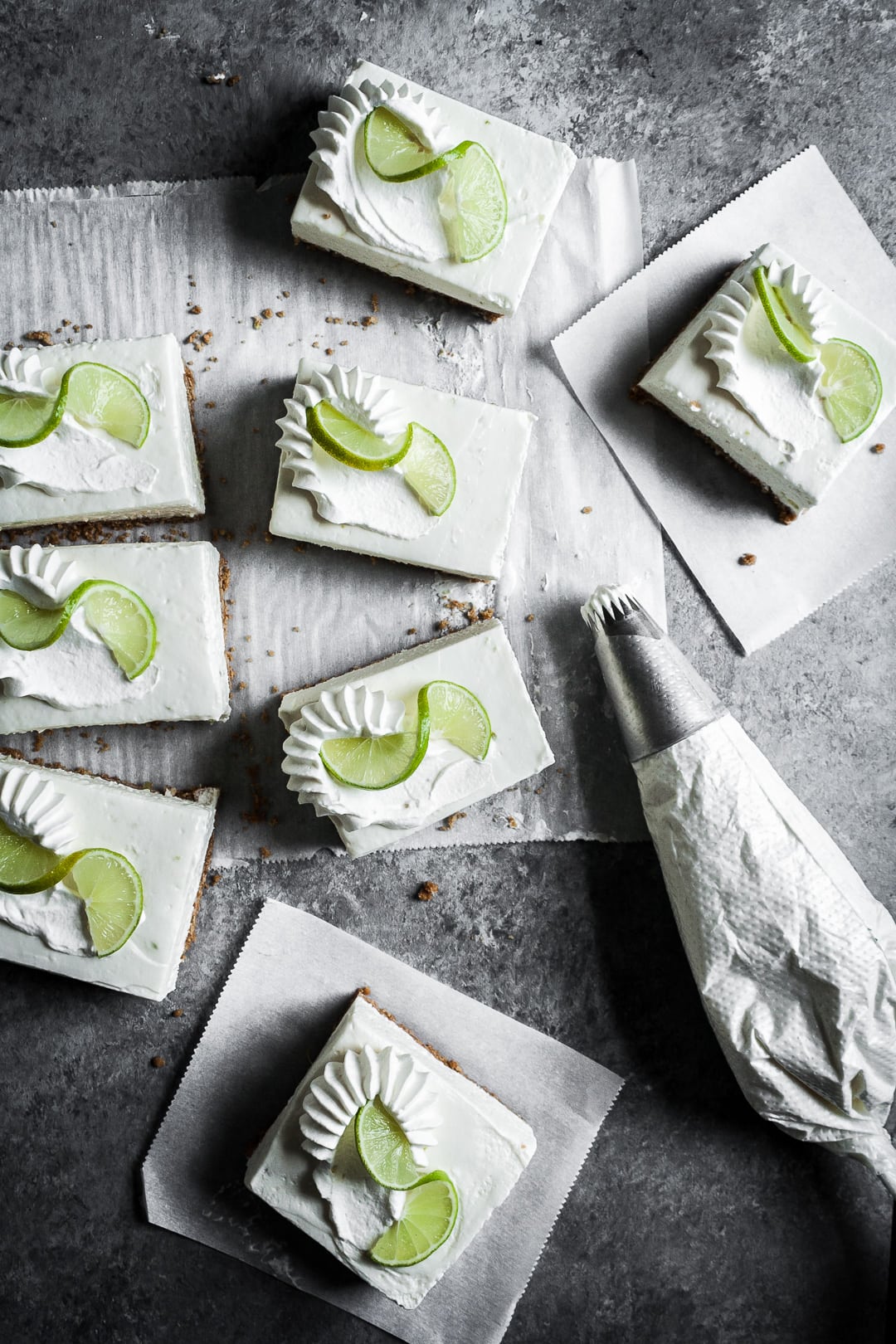 Top view of sliced cheesecake bars on parchment paper squares on a grey background with a piping bag of whipped cream nearby