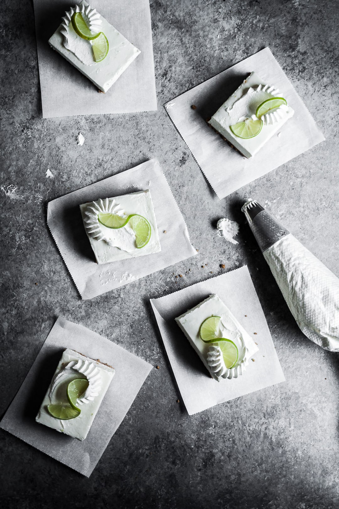 Five sliced cheesecake bars on parchment paper squares on a grey background with a piping bag of whipped cream nearby