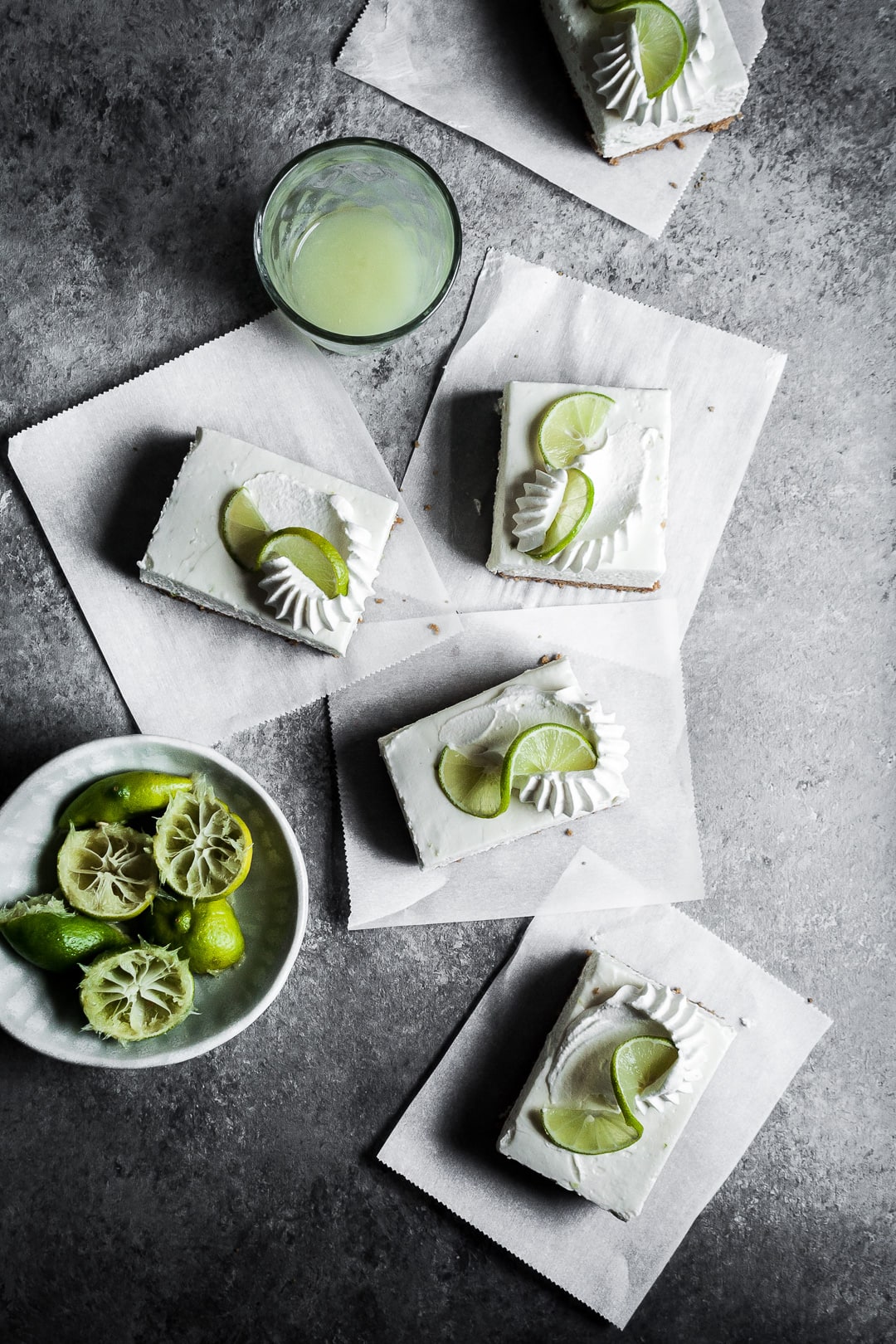 Cheesecake bar slices with piped whipped cream and lime twists on squares of parchment paper on a grey surface.