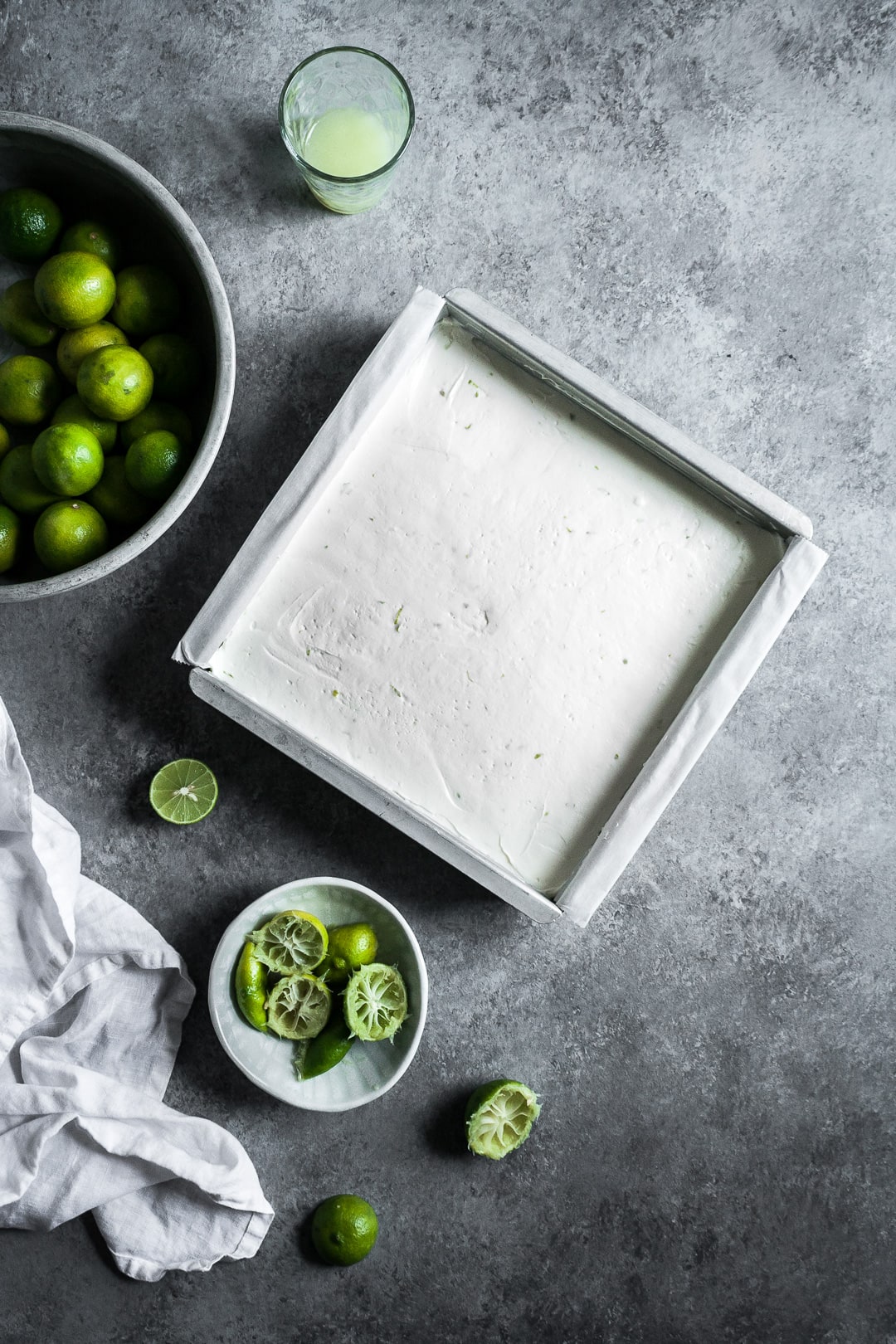 Pan of cheesecake bars on a grey background surrounded by key limes and a white linen napkin