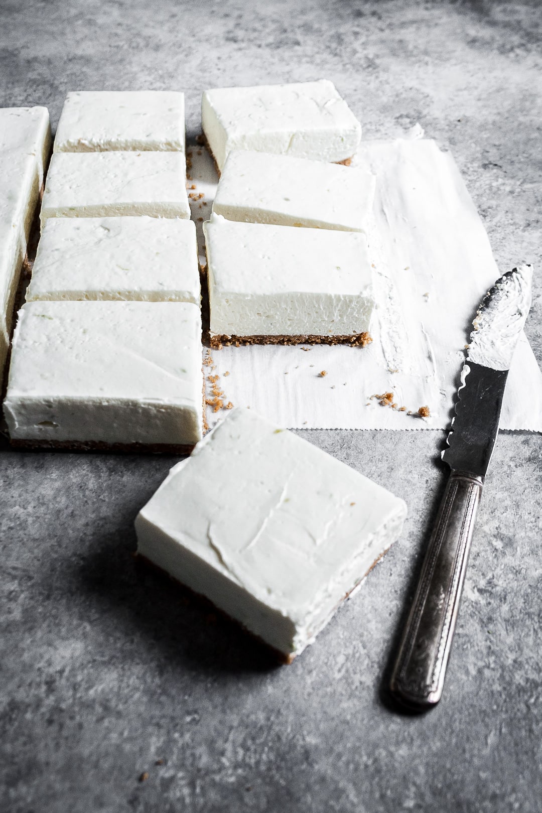 45 degree angle view of sliced cheesecake bars on parchment paper squares on a grey background with a vintage knife nearby