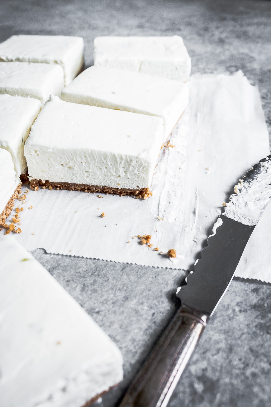 45 degree angle view of sliced cheesecake bars on parchment paper squares on a grey background with a vintage knife nearby