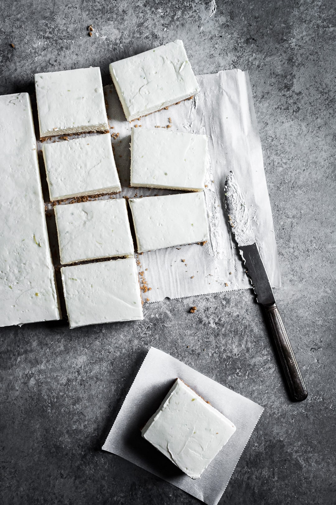 Sliced cheesecake bars on parchment paper squares on a grey background with a vintage knife nearby