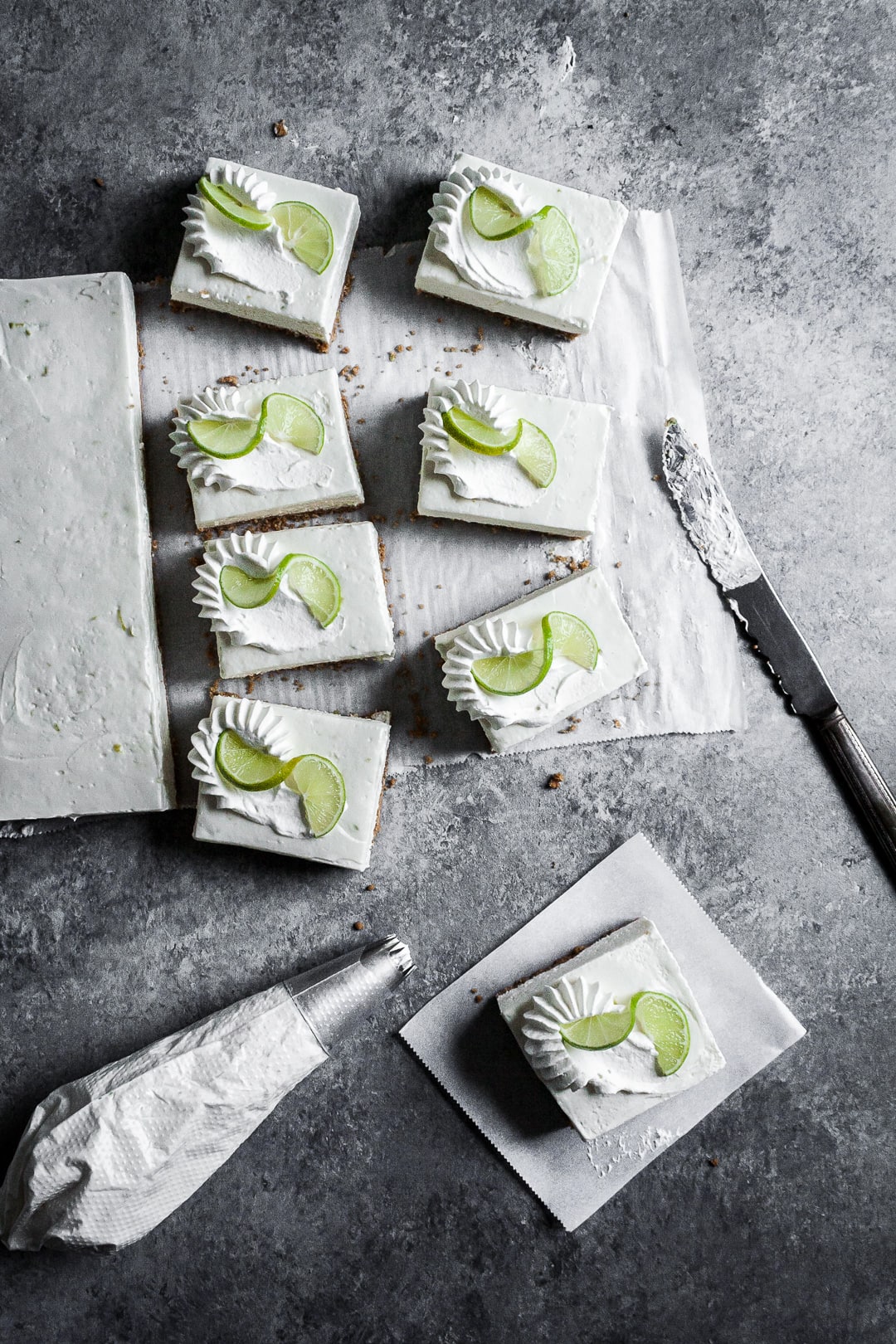 Five sliced cheesecake bars on parchment paper squares on a grey background with a piping bag of whipped cream and vintage knife nearby