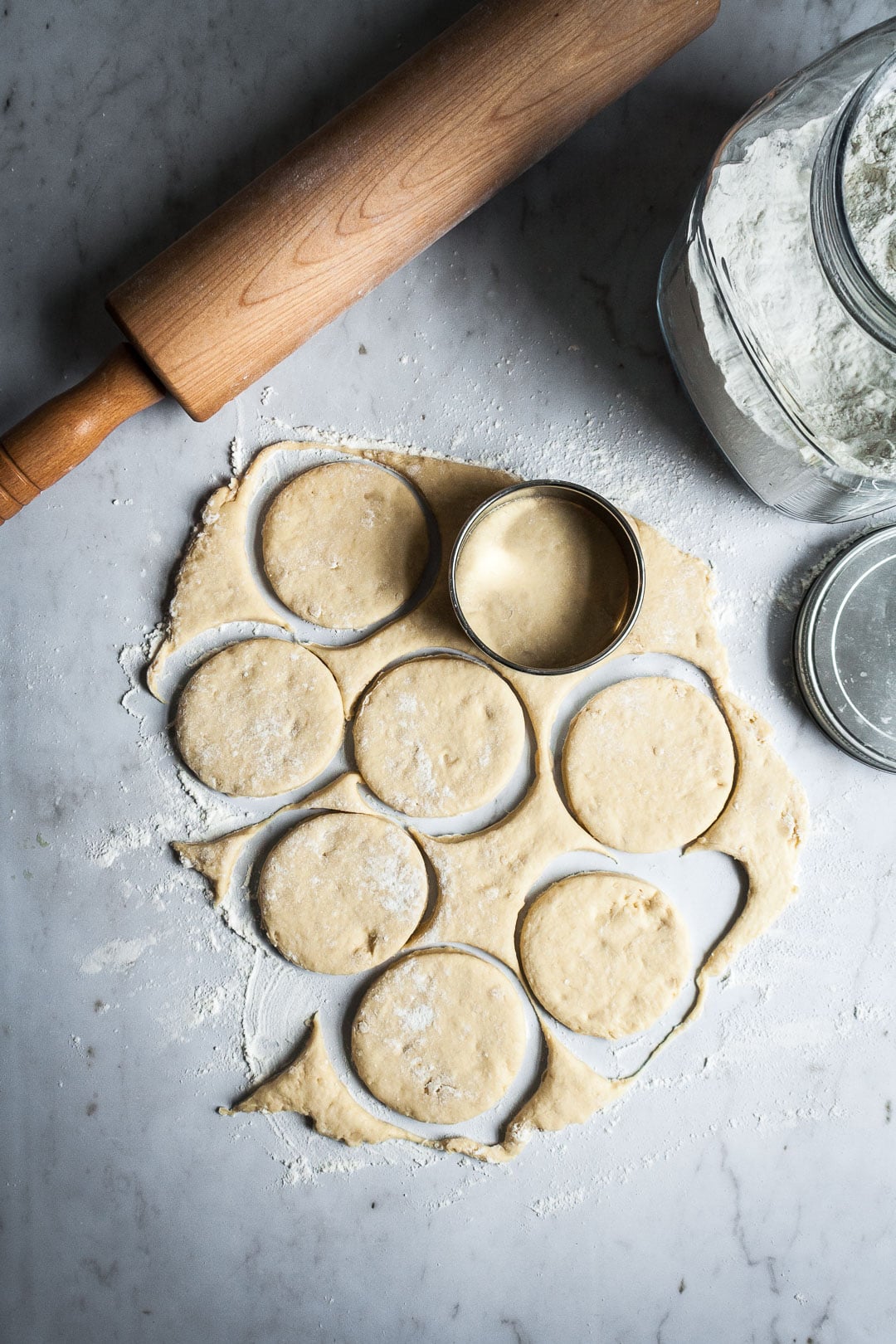 Brioche dough being rolled out and cut with a round cutter on a marble surface with rolling pin and flour container nearby