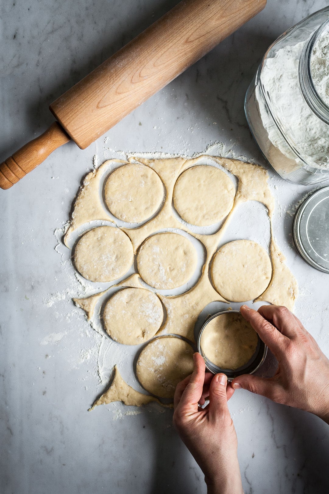 Hands cutting rolled out brioche dough into circles, holding a round cookie cutter. All on a marble slab with rolling pin and flour container nearby