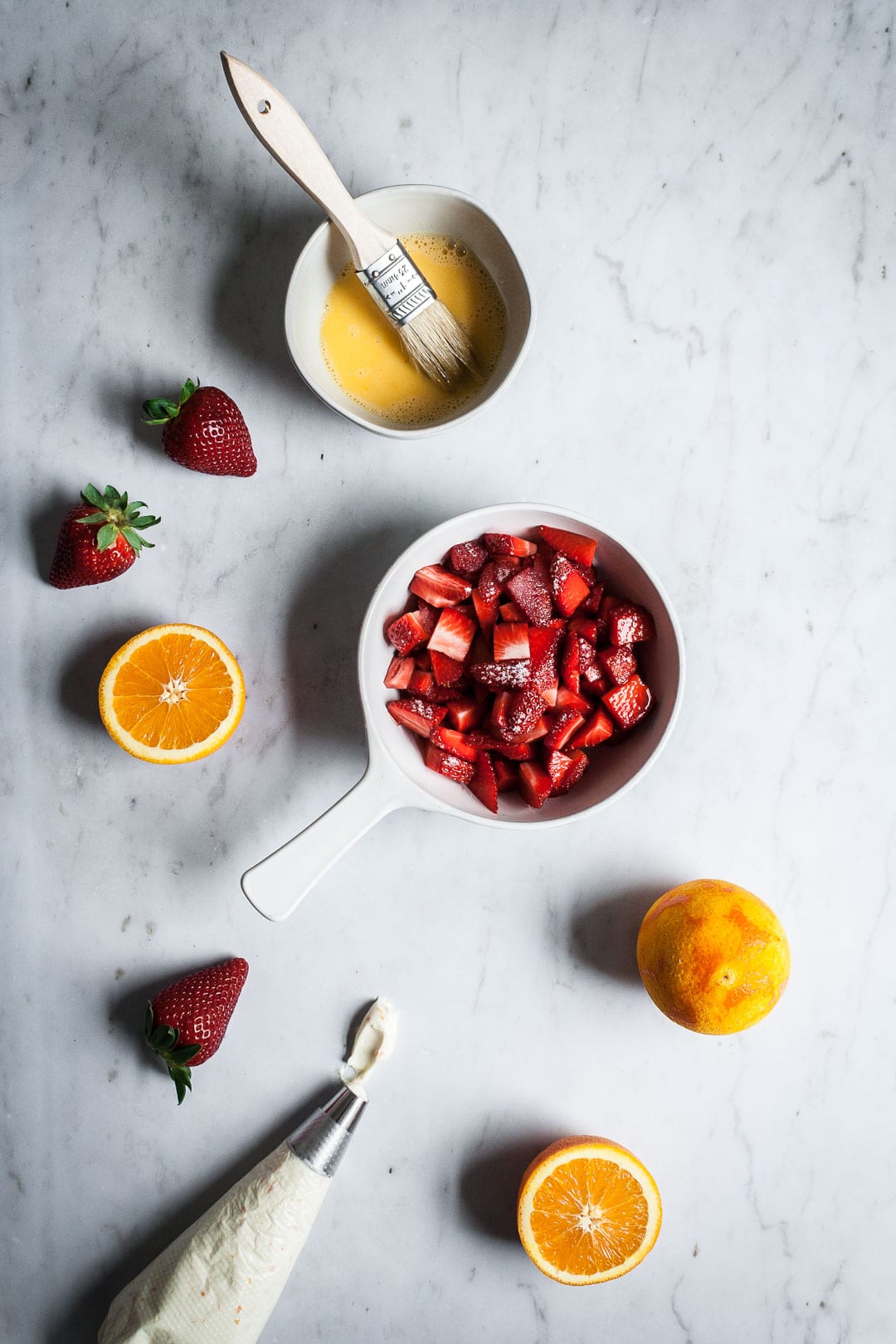 Top view of ingredients to prepare brioche pastries, including egg wash, strawberries, and cream cheese filling in piping bag