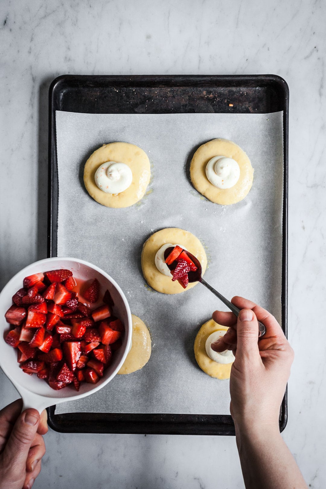 Process shot showing hands holding bowl of strawberries, spooning them onto the top of cream cheese brioche pastries