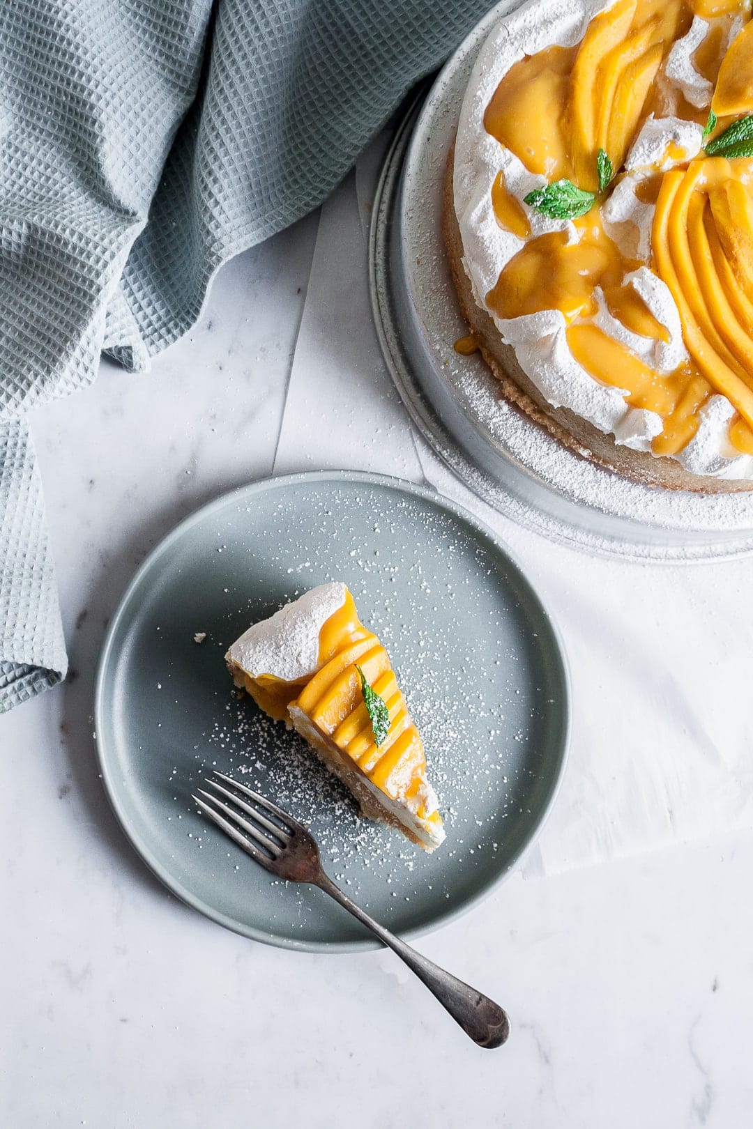 Slice of coconut meringue mango cake on a blue plate with rest of cake nearby