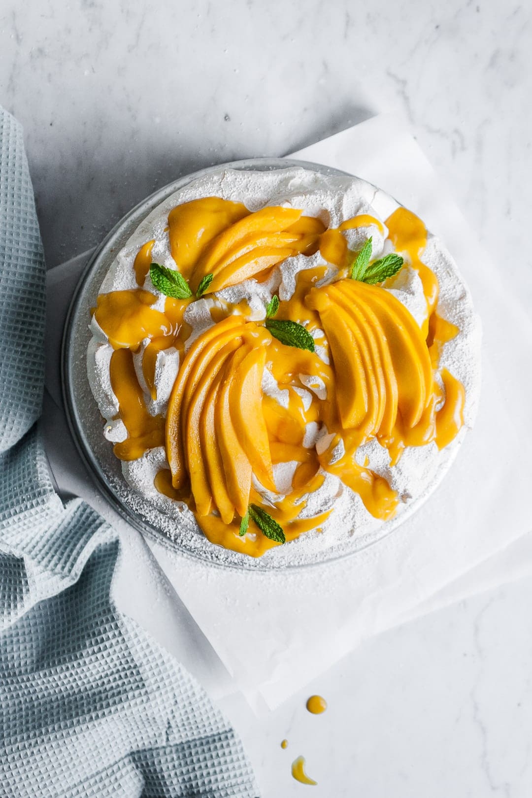 Top view of meringue cake with mango sauce and sliced mangoes on top, garnished with mint leaves on a white marble background
