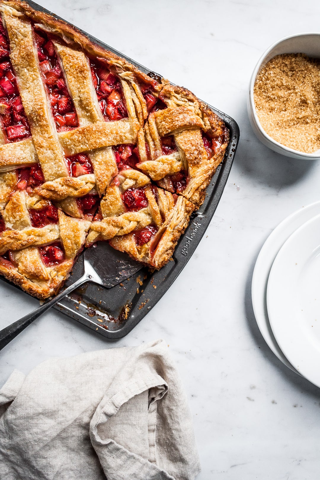 Baked strawberry rhubarb slab pie in a sheet pan with several cut slices and a serving utensil on a marble background