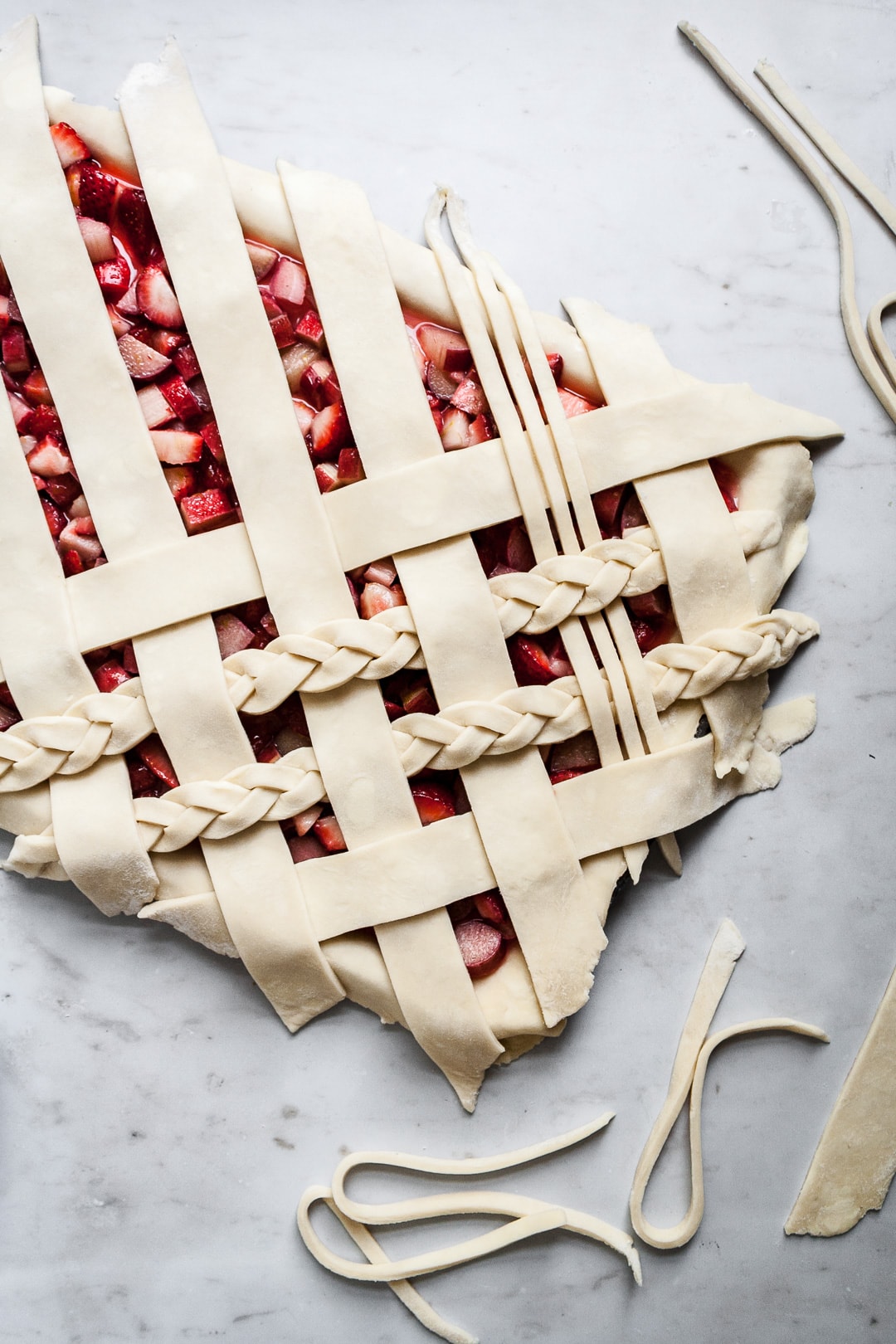 Top view of unbaked strawberry rhubarb slab pie with angled woven lattice crust with braids on a marble background with dough scraps nearby