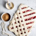 Top view of unbaked strawberry rhubarb slab pie with angled woven lattice crust with braids on a marble background and bowls of egg wash and coarse sugar nearby