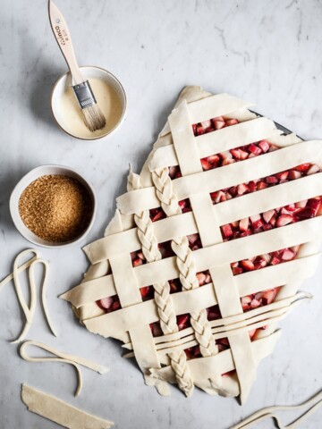 Top view of unbaked strawberry rhubarb slab pie with angled woven lattice crust with braids on a marble background and bowls of egg wash and coarse sugar nearby