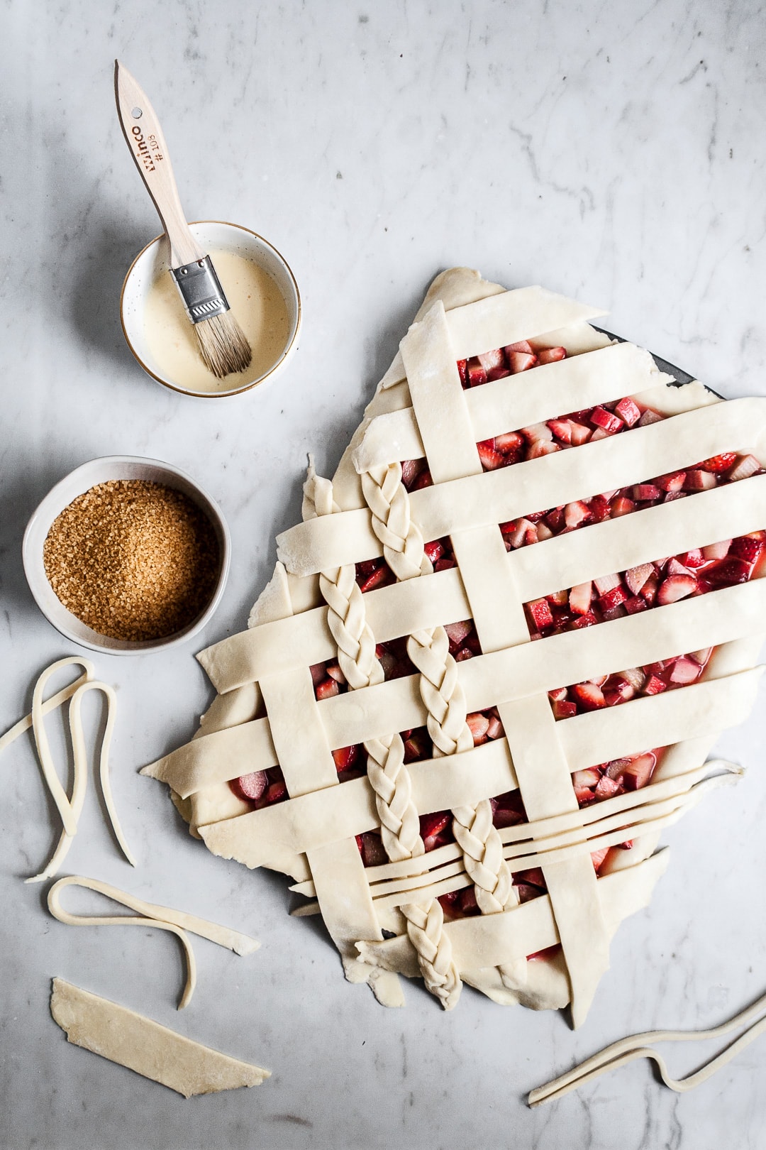 Top view of unbaked strawberry rhubarb slab pie with angled woven lattice crust with braids on a marble background and bowls of egg wash and coarse sugar nearby