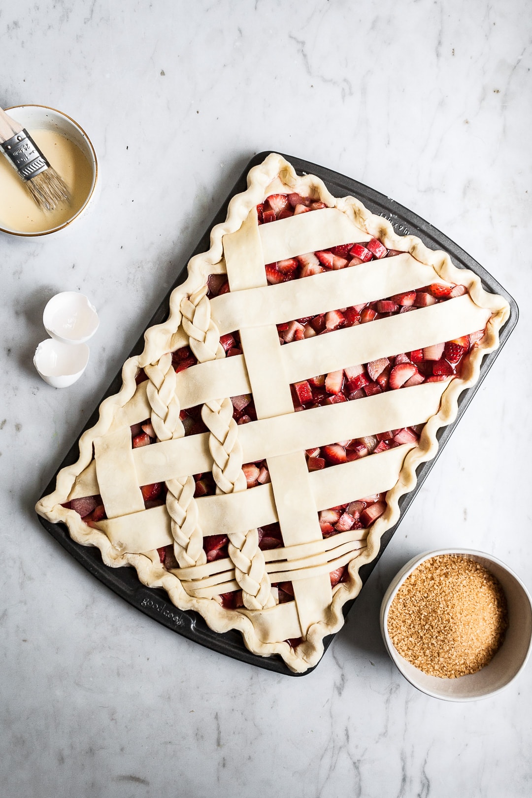 Crimped unbaked lattice slab pie crust in sheet pan on a marble background with bowls of sugar and egg wash nearby