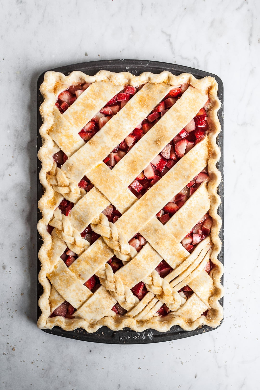 Top view of unbaked crimped lattice slab pie with strawberry rhubarb filling, sprinkled with coarse sugar, on a marble background