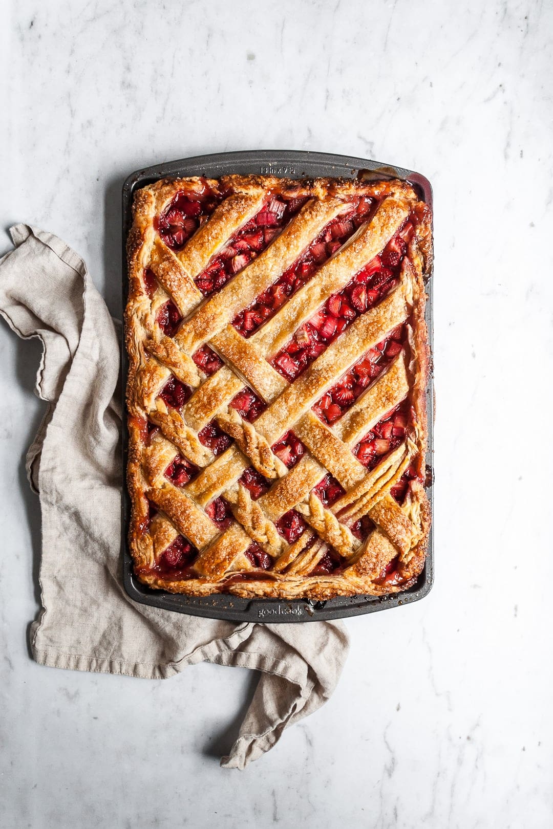 Top view of baked strawberry rhubarb slab pie with natural linen towel underneath pan, resting on a marble background