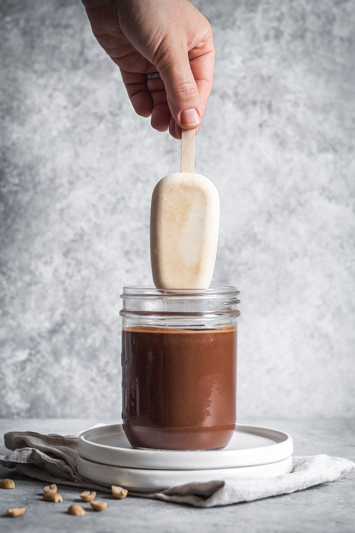 Hand dipping ice cream bar into chocolate with grey background behind