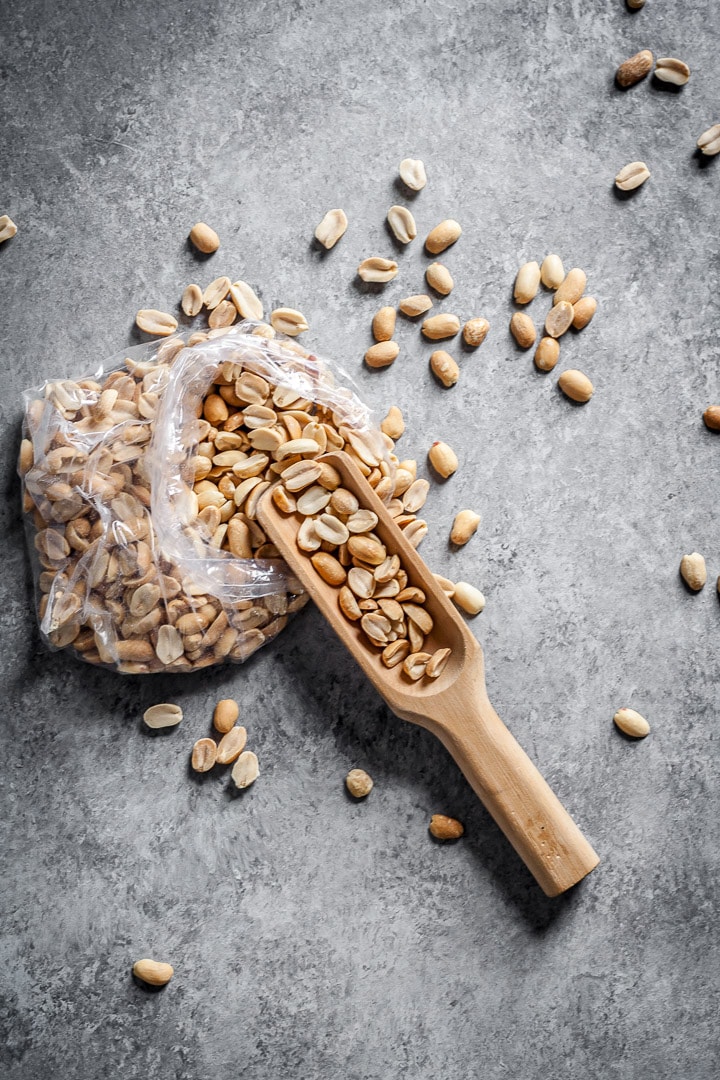 Bag of peanuts strewn on grey surface with a wooden scoop