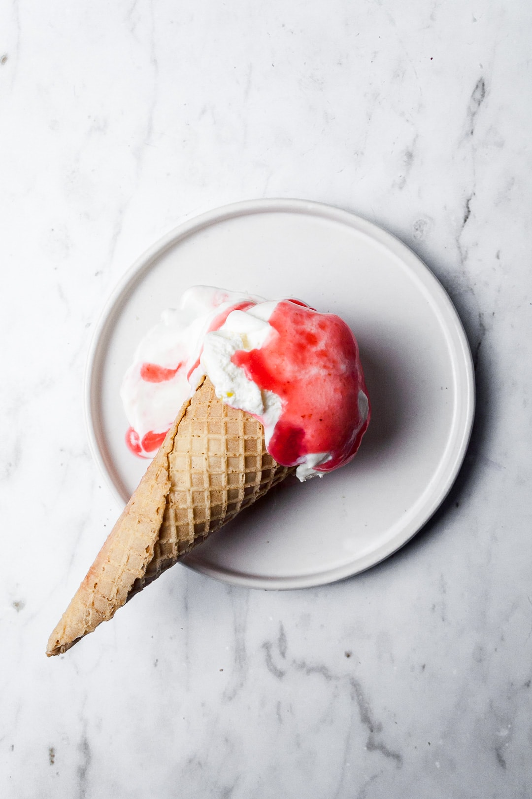 Top view of sugar cone with scoop of ice cream and drizzle of red sauce on a white plate and marble background