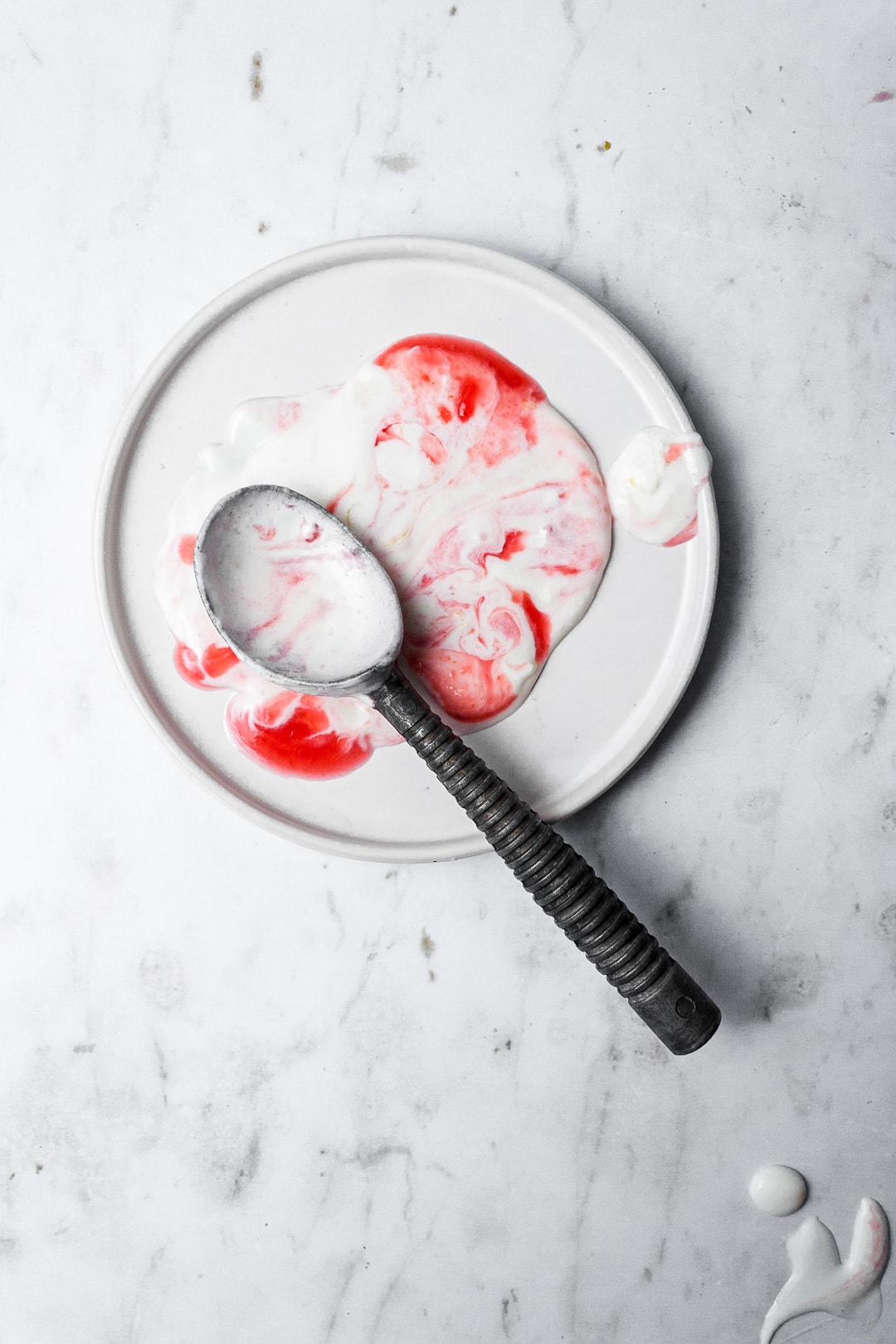 Melted puddle of marbled red and white ice cream on a white plate with a vintage ice cream scoop on top