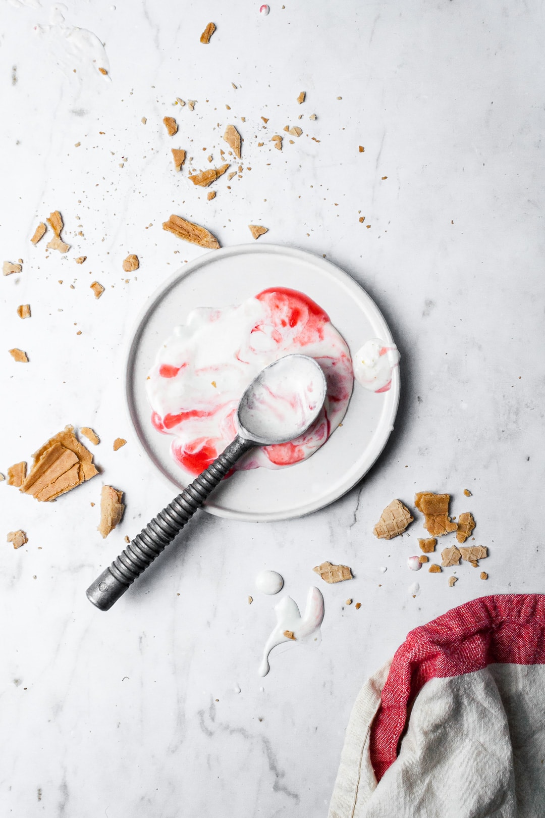 Melted puddle of red and white ice cream on a white plate with crushed sugar cone strewn nearby