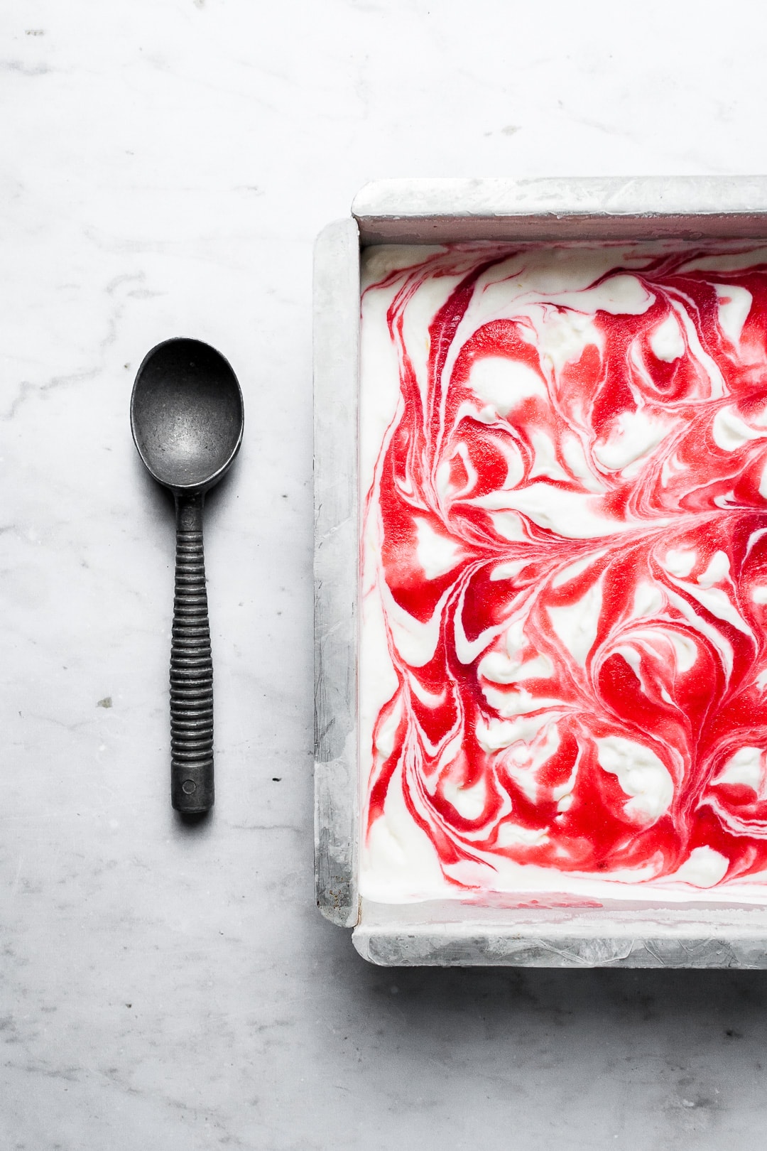 Metal pan of white ice cream with red berry swirl on a marble backdrop with a vintage ice cream scoop nearby