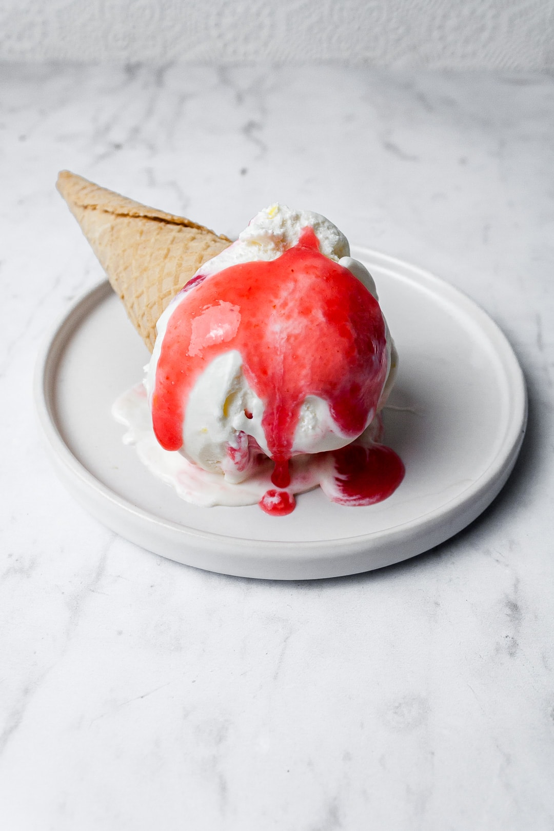 Scoop of ice cream in a sugar cone with a red sauce dripping onto a white plate and marble background