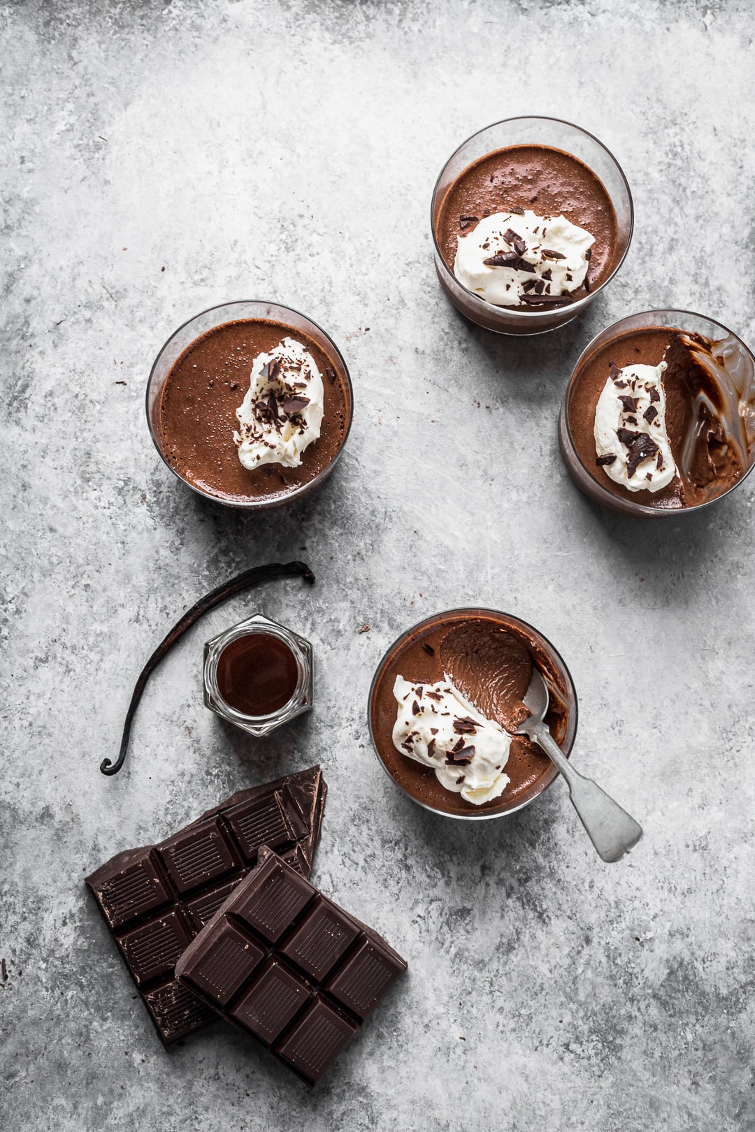 Four chocolate pots de creme on a grey background with chocolate and vanilla nearby