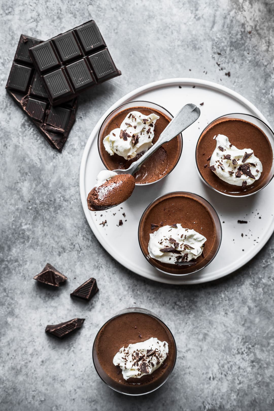 Top view of chocolate pots de creme on a white plate and grey background with chocolate scattered nearby