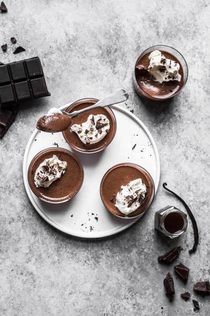 Four chocolate desserts in glasses on a white plate and grey background with chocolate and vanilla nearby