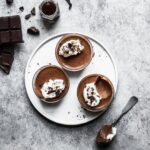 Chocolate pots de creme on a white plate and grey background with ingredients and spoon nearby