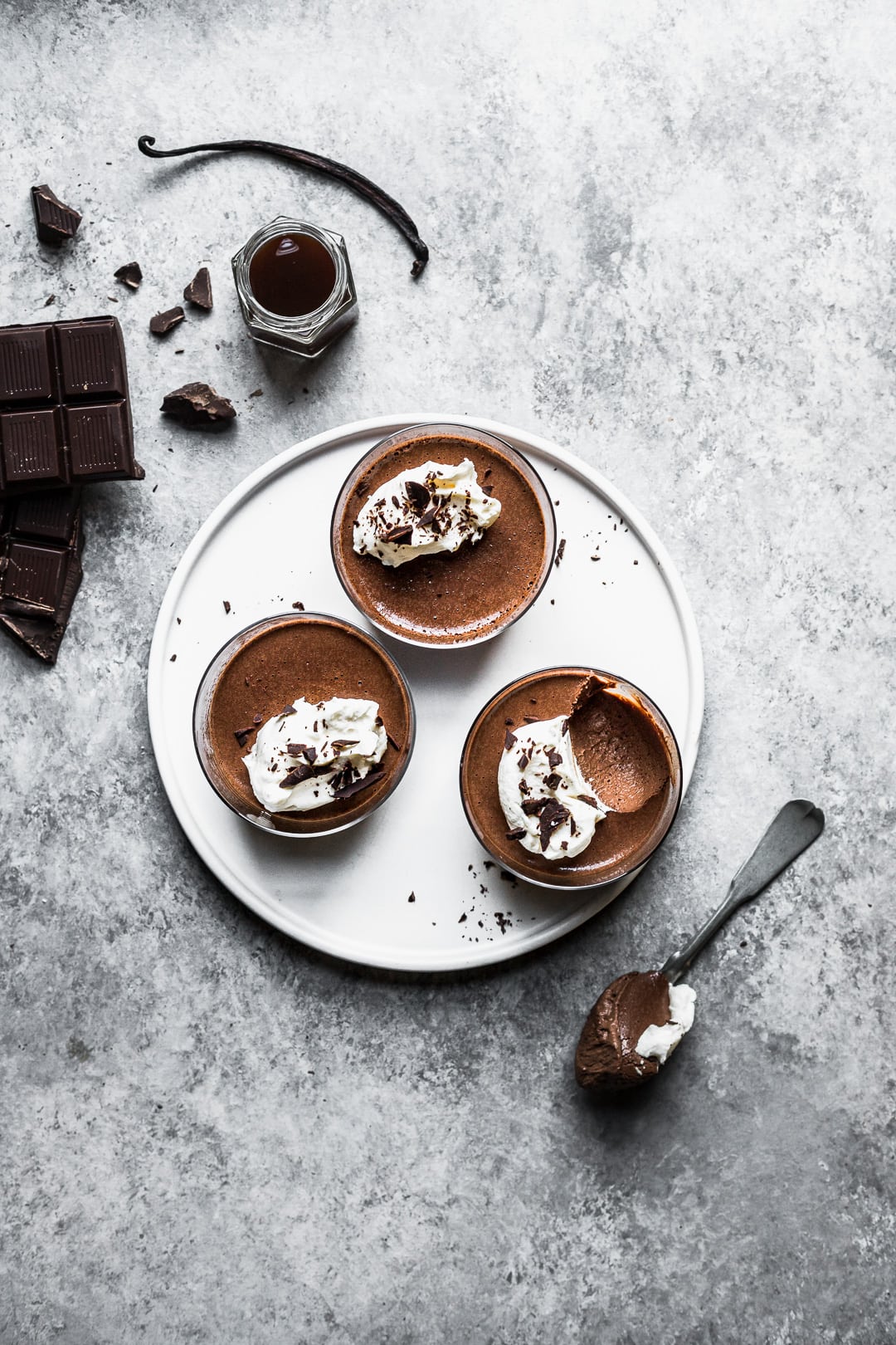 Chocolate pots de creme on a white plate and grey background with ingredients and spoon nearby