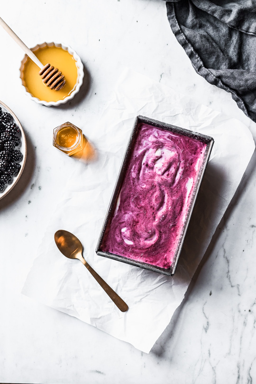 Purple and white swirled ice cream in a container on a marble background with blackberries and honey nearby