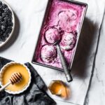 Three scoops of purple ice cream in container on a marble background with honey and blackberries nearby