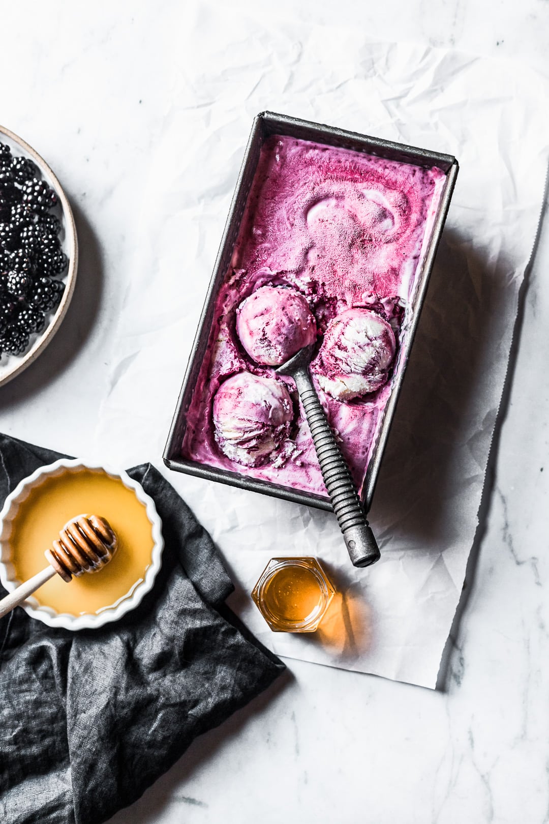 Three scoops of purple ice cream in container on a marble background with honey and blackberries nearby