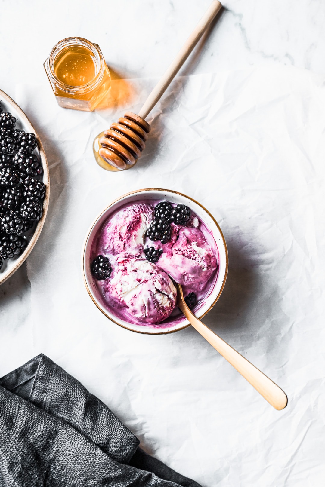 Three scoops of purple and white swirled ice cream in a bowl with honey and blackberries nearby