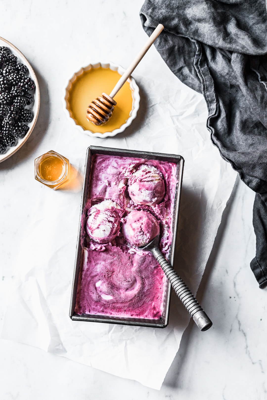 Three scoops of purple ice cream in container on a marble background with honey and blackberries nearby