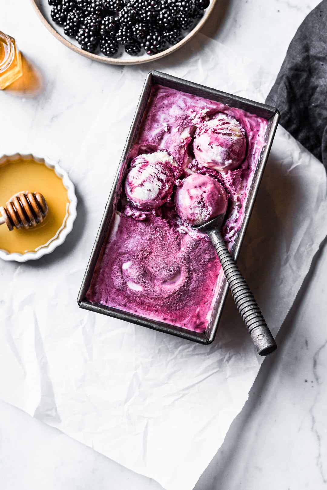 Three scoops of purple ice cream in container on a marble background with honey and blackberries nearby