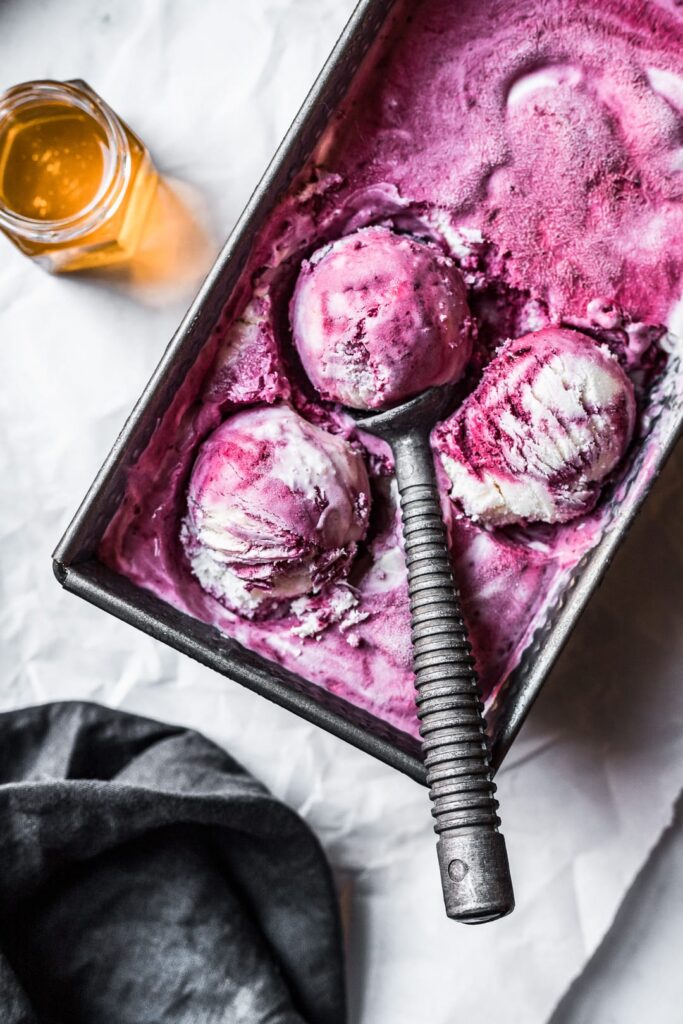 Three scoops of purple and white ice cream in a container on parchment paper with honey nearby