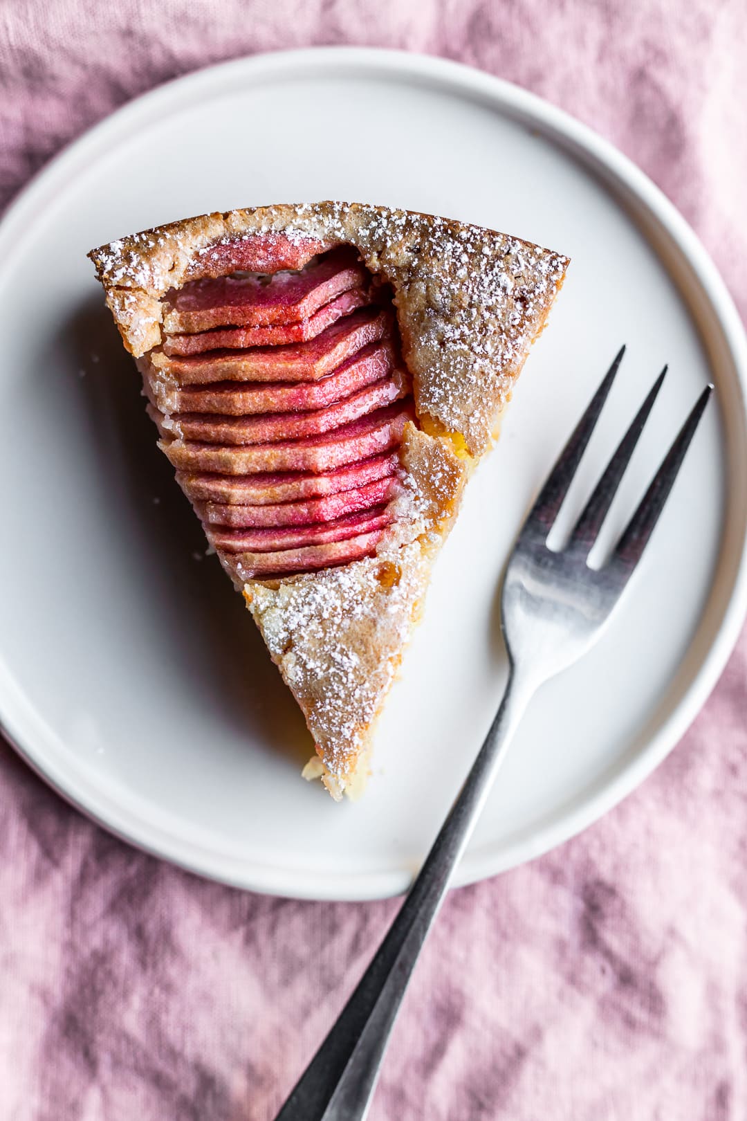 Slice of almond cake with sliced pink apples on a white plate on a pink napkin