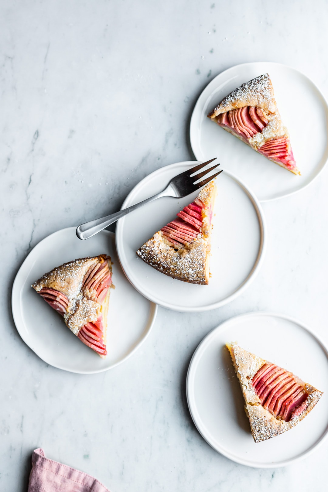 Slices of almond cake with pink apples on four white plates on a marble background