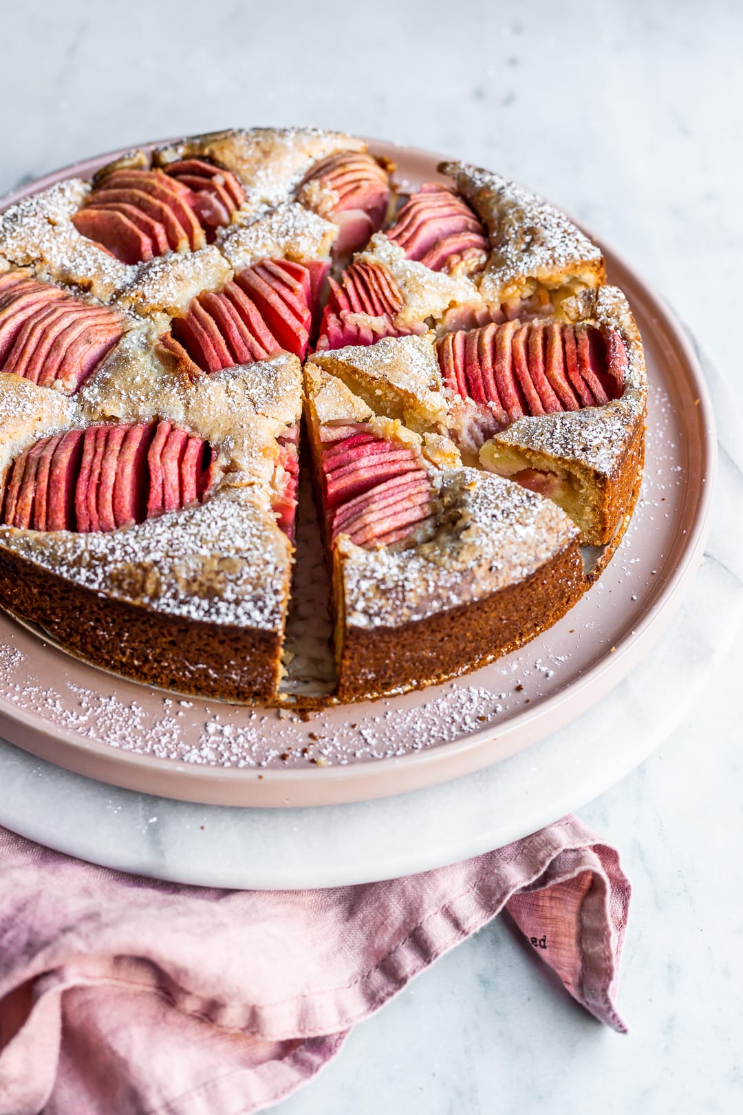 Angled view of sliced almond cake with pink apples on a pink plate and marble background